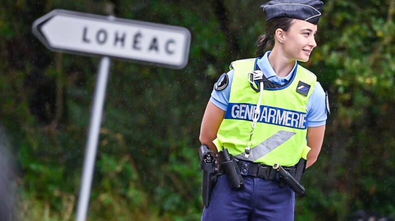 Une gendarme porteuse de chasuble jaune, regardant vers la doite et les bras dans le dos. En arrière-plan, un panneau de direction de Loheac et la forêt