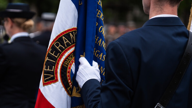 On aperçoit un militaire de dos, en uniforme bleu marine, tenant dans sa main droite gantée un drapeau tricole