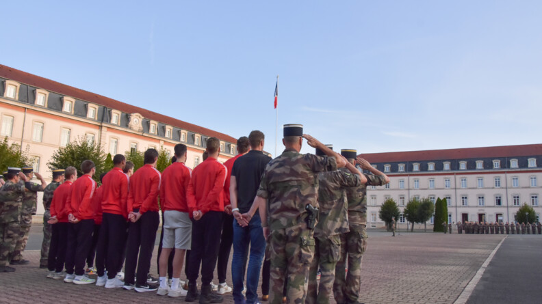 Au premier plan, sur la place d'armes, vue de dos des sportifs en tenue de sport et des militaires de la gendarmerie en treillis kaki, au garde-à-vous. De l'autre côté de la place d'armes, une compagnie d'élèves gendarmes au garde-à-vous. Au fond et à gauche, les bâtiments de l'école.