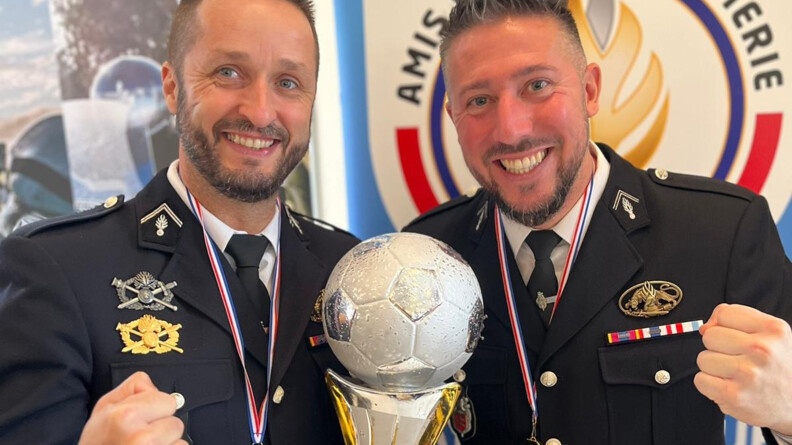 Deux militaires souriants, en blazer sombre, posent souriants devant un troph&eacute; de futsal, que l'un d'eux tient dans la main gauche. Tous deeux portent une m&eacute;daille dor&eacute;e autour du cou, et tiennent leur poing serr&eacute; en signe de victoire