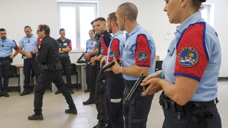 Des élèves en ligne, pistolet à la main regarde l'instructeur qui montre une position de tir