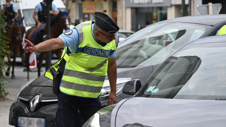 Un gendarme, revêtant un calot noir et une chasuble jaune, s'adresse à un automobiliste, la tête penchée dans sa direction, et désignant un point avec l'index de sa main droite.