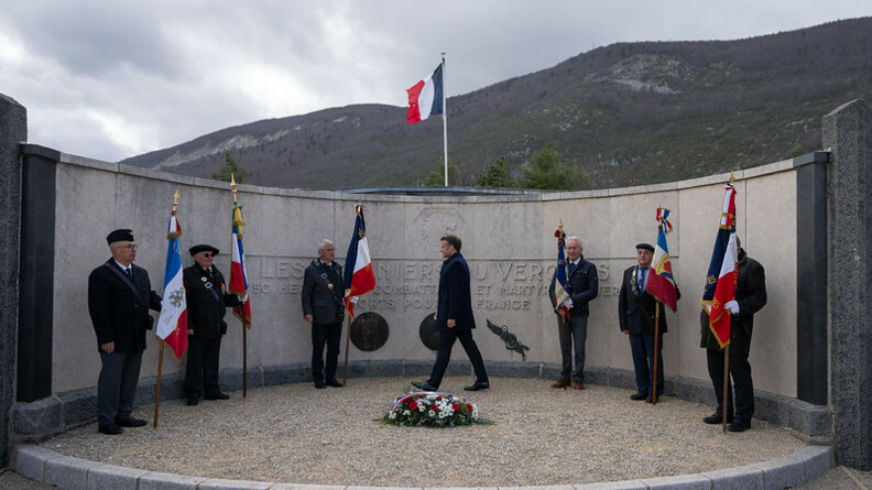 Agrandir l'image Devant un monument circulaire en pierre, le chef de l'Etat, Emmanuel Macron, marche devant des anciens combattants ou amis de la Résistance, se tenant debout, tenant le manche d'un drapeau dans leur main. Au sol est posée une gerbe de fleurs recouvrte d'une bannière bleue, blanc, rouge.