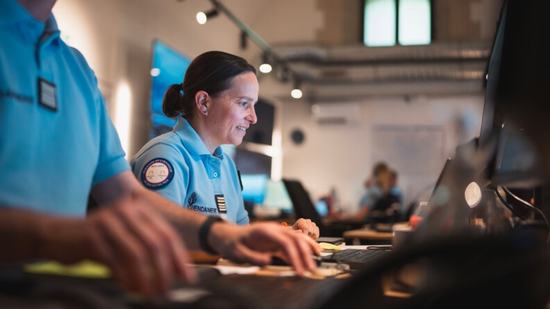 Une femme gendarme travaille sur son ordinateur, posé sur une table. Elle apparait de biais sur la photo, aux côtés d'un autre gendarme, dont on distingue le torse et les mains, travaillant lui-aussi  sur son ordinateur.