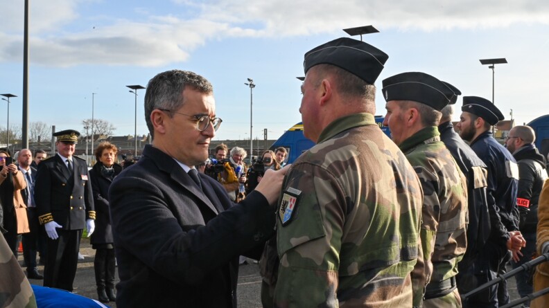 G&eacute;rald Darmanin, debout, de biais,appose sur la poitrine d'un militaire en tenue kakie une d&eacute;coration, que l'on devine mais que l'on ne voit pas. Ils sont en ext&eacute;rieur, entour&eacute;s d'hommes et de femmes, militaires, policiers et civils..