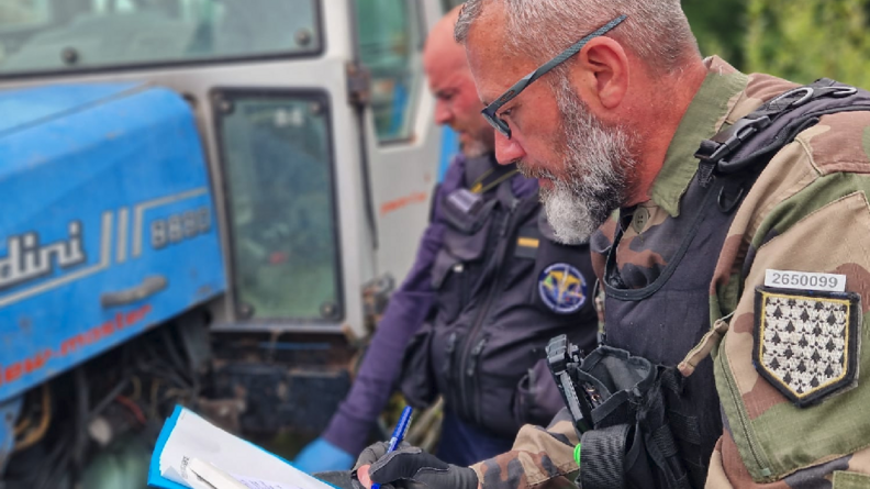 Deux gendarmes, accroupis à côté d'un tracteur, relèvent des informations. L'un d'eux prend des notes sur un calpin.