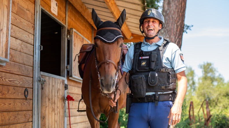 Agrandir l'image Devant le box, le cheval à gauche, le cavalier à droite, tenant les rênes sous la gorge du cheval. Debout, tout équipé avec son gilet pare-balle et sa bombe d'équitation, il rit aux éclats