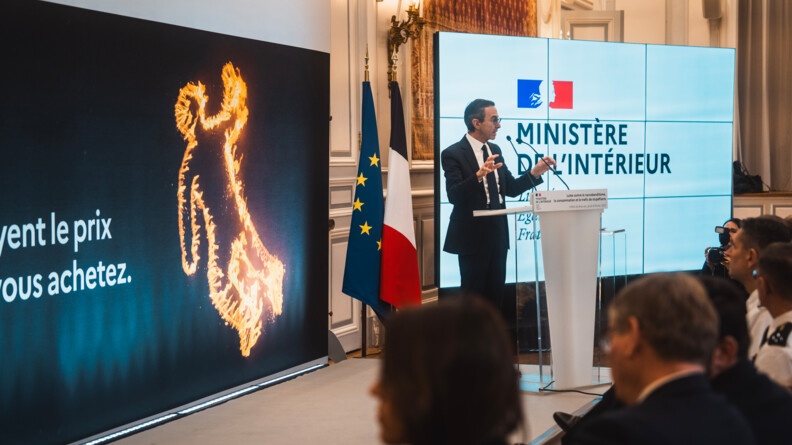 Agrandir l'image à gauche une silhouette aux contours en flammés sur fondnoir, à droite le minsitre de l'Intérieur, Bruno Retailluea, derrière un pupitre et deux micro, devnat le drpeau français et le drapeau européen et un écran géant portantla mention "Minsitère de l'Intérieur", avec tout à droite et enbas quelques têtes du public présent