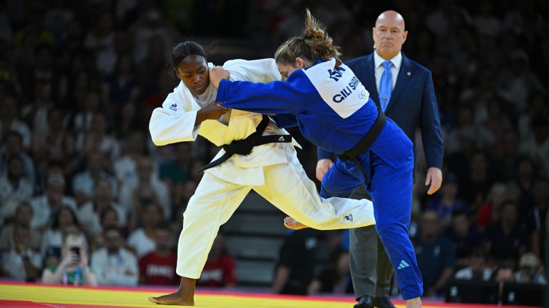 La judoka Clarisse Agbegnenou en kimono blanc lors d'un combat avec une adversaire en kimono bleu, sur un tatami rouge et jaune, sous l'oeil d'un arbitre en costume au second plan.