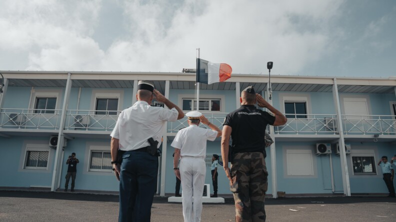 Deux gendarmes et un préfet de dos saluant le drapuea français devant un bâtiment à la façade bleu ciel et blanc