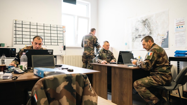 Agrandir l'image Dans une salle aux murs blancs, quatre bureaux se font face, deux par deux, avec des ordinateurs portables et du matériel de bureau. Les deux bureaux de droite et celui du fond à gauche sont occupés par des prévôts, travaillant. Au fond à droite, derrière son camarade, un prévôt de dos, debout, en train de faire couler le café. Le mur de droite arbore une carte de la Roumanie et de la documentation, le mur du fond un tableau blanc magnétique du service mensuel