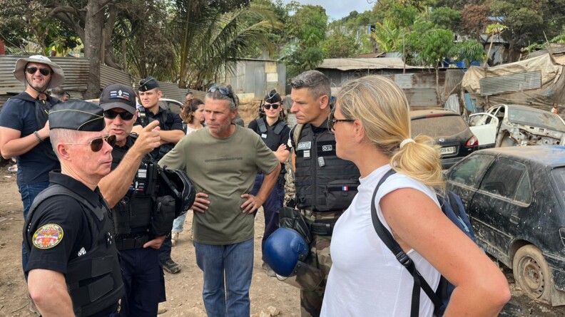 Au premier plan, le major g&eacute;n&eacute;ral de profil, main sur la hanche, face &agrave; une femme blonde, portant un tee-shirt blanc, &eacute;galement de profil et mains sur les hanches. Au second plan, vus de face, deux autres officiers de gendarmerie et un homme en civil, portant un jean et un tee-shirt kaki. Derri&egrave;re eux, deux autres officiers  et deux personnes civiles. A droite de l'image, trois &eacute;paves de voiture et dans le fond, sous les palmiers, des habitats en t&ocirc;le.