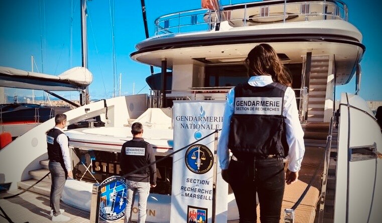 Trois gendarmes de dos, en civil, portant une chasuble bleu marine avec la mention "GENDARMERIE SECTION DE RECHERCHES" dans le dos. Ils sont debout sur le pont arrière d'un yacht sur lequel est posé un kakémono de la section de recherches de Marseille.