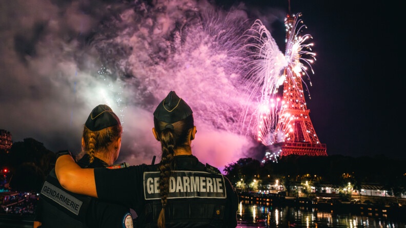 Deux femmes gendarmes, vêtues d'une tenue noire, et portant une tresse, se tiennent côte à côte, tournant le dos à l'objectif, et se tenant en bordure de Seine, face à la Tour Eiffel, devant laquelle un feu d'articice est tiré. La femme de droite a passé sa main sur l'épaule de la collègue.