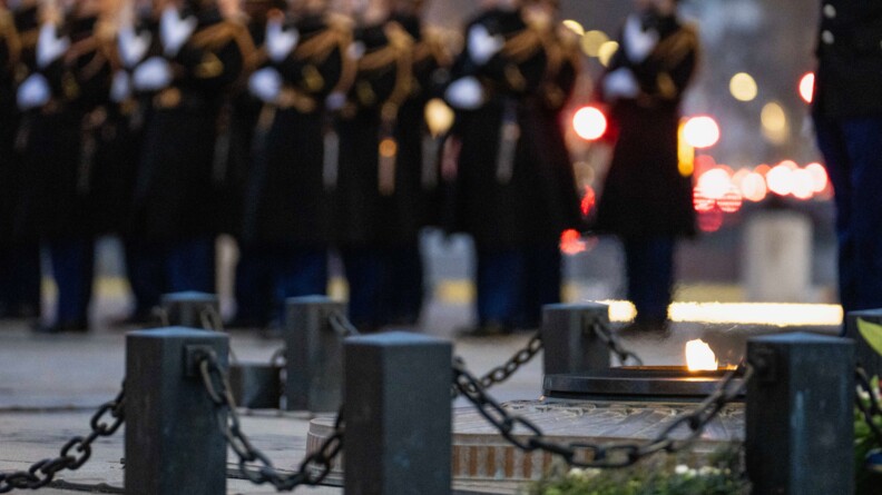 Gros plan sur la flamme du Soldat inconnu, prot&eacute;g&eacute;e par des plots et des cha&icirc;nes. En arri&egrave;re plan, en flout&eacute;, la troupe des gardes r&eacute;publicains.