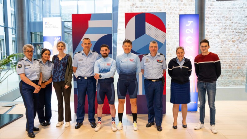 Agrandir l'image Devant un mur en pierre et des kakémonos à l'éfficgie des juex olympiques de Paris 2024, deux cyclistes se tiennent debout, souriants, entourés de sept personnels de la gendarmerie nationale. Cinq d'entre eux sont en tenue de gendarme, deux autres en tenue civile.
