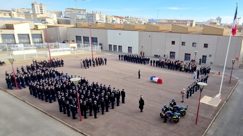 Vue a&eacute;rienne d'une place d'armes, au milieu de b&acirc;timents de couleur beige. Au milieu, repose un cercueil recouvert du drapeau tricolore. A gauche, trois pelotons de gendarmes sur huit rang&eacute;es, au garde-&agrave;-vous. Au bout de la place, deux groupes de personnes en civil. A droite, un groupe de militaires en tenue et un groupe de personnes en civil, ainsi qu'un g&eacute;n&eacute;ral derri&egrave;re un pupitre.