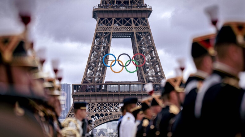 photo de la Tour Eiffel revêtant les anneaux olympiques. Devant elle, de biais, se tiennent des gardes républicains, dont l'image est floutée.