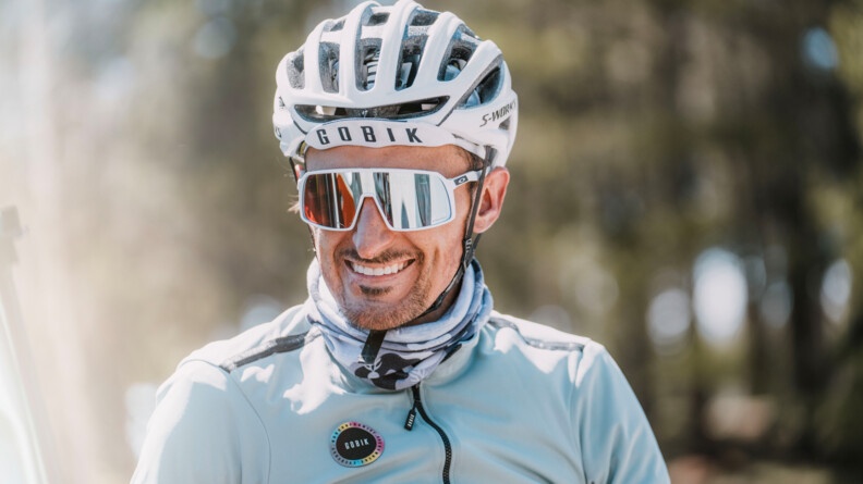Un cycliste, dont on aper&ccedil;oit uniquement le haut du corps, regarde l'objectif en souriant. Il es v&ecirc;tu d'un blouson bleu ciel, d'un casque blanc et de lunettes de soleil blanches.