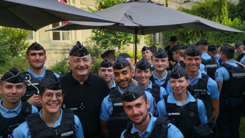 Une quinzaine d'élèves-gendarmes de Dijon autour du directeur général de la gendarmerie nationale, dans les jardins du ministère de l'Intérieur. En arrière-plan, d'autres gendarmes de dos, des parasols beiges, de la verdure et des bâtiments.