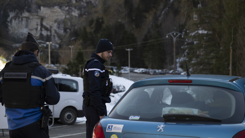Deux gendarmes, celui de gauche de dos, celui au centre de profil, abordent un véhicule peugeot bleu ciel dans une route à lacets aux pieds de montagnes