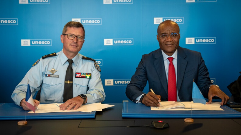 Agrandir l'image A gauche un général de gendarmerie et à droite un homme en civil s'apprêtent à signer une convention. Au fond, un mur bleu portant à de nombreuses reprise le logo de l'UNESCO