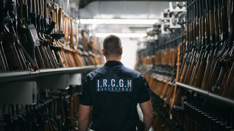 Au centre de l'image, un homme apparait de dos. Il porte un uniforme bleu marine, à manches courtes, avec dans le dos l'inscription "I.R.C.G.N Balistique", ainsi qu'une arme à la ceinture. l'homm est châtain et ses cheveux sont coupés court. Il circule dans une allée étroite, dans lesquelles sont accrochés de nombreux fusils.