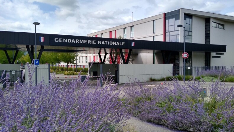 Photo de l'entrée d'une caserne de gendarmerie, et d'un bâtiment que l'on aperçoit au-delà du porche. En premier plan, au niveau de l'entrée, des lavandes sont plantées au sol.