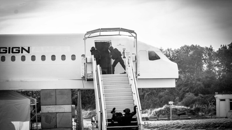 Agrandir l'image Trois hommes du GIGN au moment d'un assaut sur une maquette de carlingue d'avion. Deux sont debout, de dos, à la porte de l'avion. Le troisième dégringole l'escalier menant à la porte