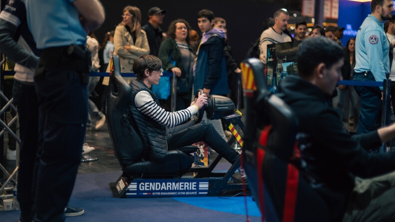 Dans le hall du salon, au milieu de nombreux visiteurs, on voit un adolescent assis dans un simulateur de conduite, au bas duquel est inscrit Gendarmerie. Ses deux mains sont positionnées sur le volant. Il regarde l'écran se trouvant devant lui sur lequel on aperçoit l'intérieur d'une voiture en mouvement.