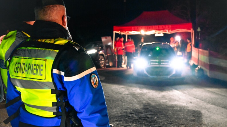 De nuit et en extérieur, un gendarme regarde un véhicule partir.