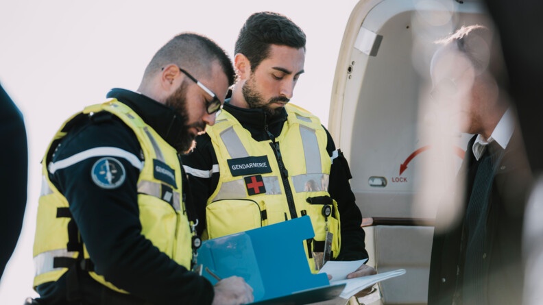 Deux gendarmes de la brigade des transports aériens, revếtant un gilet chasuble jaune au-dessus de leur uniforme bleu marine, contrôlent le commandant de bord d'un avion d'affaires. Les deux hommes tiennent en main des documents. L'un d'eux remplit un papier positionné sur un support rigide bleu. On distingue la porte de l'aéronef restée ouverte.