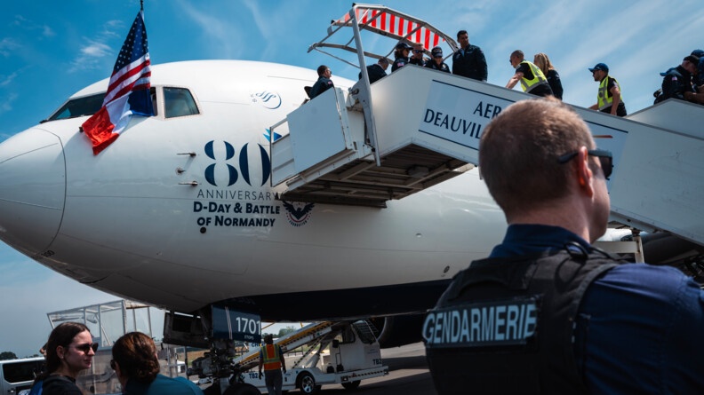 Vue en contre-plongée sur la descente d'escalier à la sortie d'un avion, d'un vétéran. Entouré de persones l'aidant (gendarmes, américains). l'avion est blanc et siglé aux 80 ans du débarquement. Au premier plan tout à droit, buste de dos d'un gendarme