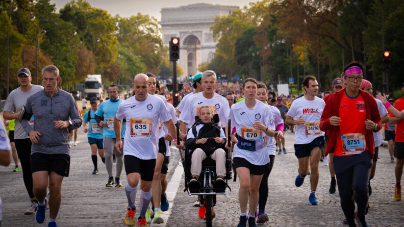 Des coureurs, vêtus d'un T-shirt blanc avec un numéro sur le ventre, courent sur une avenue pavée, en poussant le fauteuil roulant d'une jeune femme blonde, souriante, portant des tresses. On aperçoit l'arc de triomphe en arrière plan.