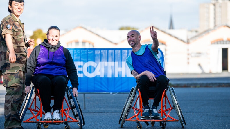 Deux personnes dans des fauteuils roulants rouge, devant une grille surmontée d'un panneau décathlon. l'homme de droite fait le sign eV de sa main gauche. A gauche de l'image, une gendarme en treillis sourit