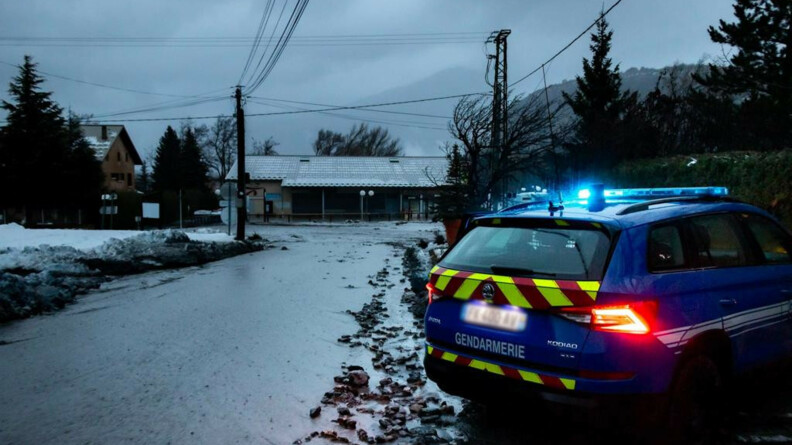 Un véhicule gendarmerie, vu de trois-quarts arrière, gyrophare allumé, stationné en bordure d'un chemin inondé, à la nuit tombante. En arrière-plan, un fleuve d'eau boueuse dévale au milieu des habitations.