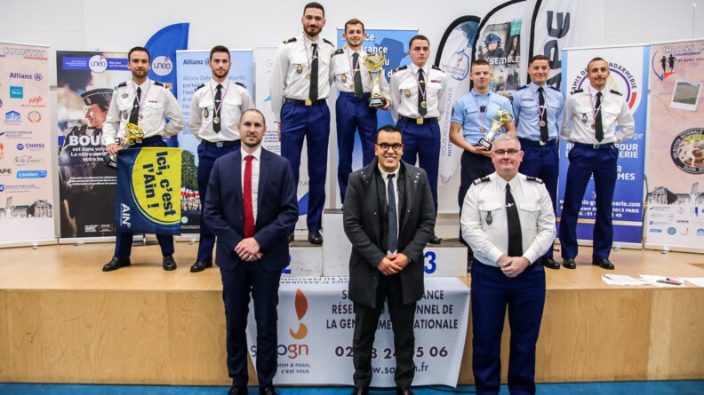 Agrandir l'image Sur la scène et le podium, huit coureurs en tenue chemise blanche et pantalon bleu sauf deux en tenue d'intervention, avec leurs médailles et coupes. Au premier plan au pied de la scène trois hommes en civil sauf un à droite, colonel de gendarmerie en tenue chemise blanche