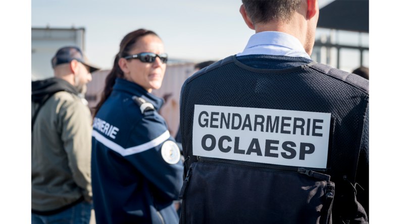 Deux gendarmes, l'un de dos et l'une de côté en flou. Celui de dos porte une chassuble avec noté dessus : Gendarmerie OCLAESP