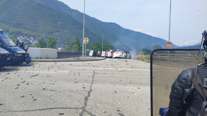 Agrandir l'image Au premier plan, à droite, un gendarme mobile vu de dos, portant un bouclier. A gauche, l'avant de deux Irisbus et plusieurs gendarmes mobiles. Face à eux, sur la route, laquelle est jonchée de pierres et autres projectiles, un groupe de manifestants radicaux.