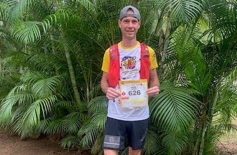 Agrandir l'image Le gendarme Maël, vêtu d'un short noir et d'un maillot blanc avec les manches et le col jaunes, portant le logo du Grand Raid, pose devant des palmiers, tenant son dossard numéro 626 entre les mains.