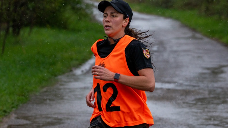 Photo d'une femme, v&ecirc;tue d'un t-shirt noir, d'une chasuble orange et d'une casquette noire, courant sur un chemin par un temps pluvieux.