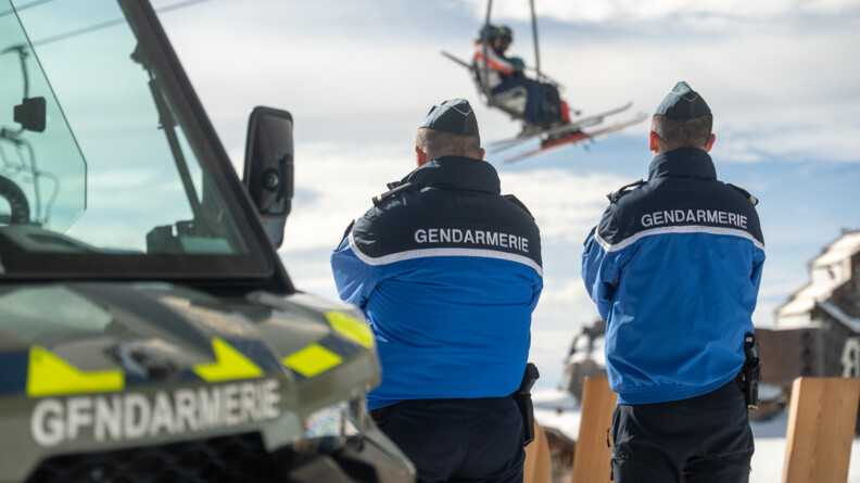 Agrandir l'image Deux gendarmes se tiennent debout, côte à côte, de dos, à côté du traxter. Les deux militaires sont tournés vers un télésiège transportant des skieurs.