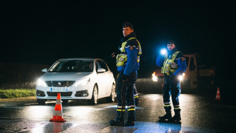 Agrandir l'image Deux gendarmes sur la droite contrôlant des voitures. On peut voir une voiture blanche à gauche de l'image.
