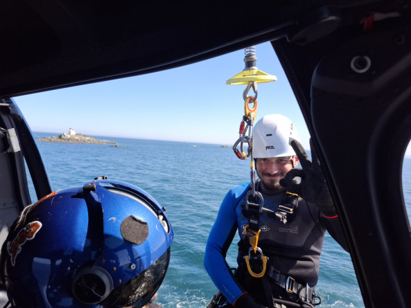 Un plongeur en combinaison n&eacute;opr&egrave;ne noire et bleue, &eacute;quip&eacute; d&rsquo;un casque blanc et d&rsquo;un harnais, est suspendu &agrave; un c&acirc;ble de treuillage en bordure d&rsquo;un h&eacute;licopt&egrave;re au-dessus de la mer. Il sourit et fait un signe "OK" avec la main droite. Un autre membre de l&rsquo;&eacute;quipage, portant un casque bleu brillant, est visible au premier plan &agrave; l&rsquo;int&eacute;rieur de l&rsquo;h&eacute;licopt&egrave;re. &Agrave; l&rsquo;arri&egrave;re-plan, on aper&ccedil;oit un petit &icirc;lot rocheux sur lequel se trouve un phare blanc.
