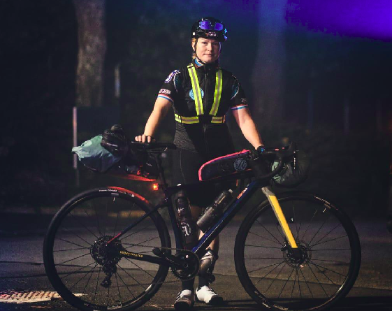 Agrandir l'image Une jeune femme cycliste pose de nuit devant son vélo. Elle se tient debout, une main posée sur la selle, l'autre sur le guidon de son vélo. Elle porte un casque et des lunettes par-dessus au niveau du front.