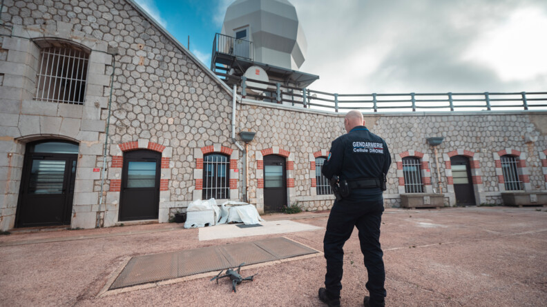 Dans un fort sécurisé, un gendarme de l'armement se prépare à faire voler son drone pour surveiller les environs. Sur son dos, est inscrit : Gendarmerie Cellule Drone