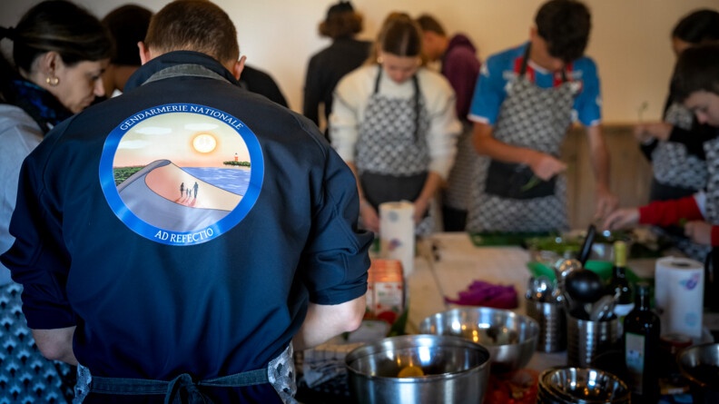 Au premier plan, de dos, un homme portant un sweat-shirt bleu marine avec une rondache dans le dos "Gendarmerie nationale - Ad Refectio". Il s'affaire devant une table où l'on voit des saladiers, des ingrédients et d'autres ustensiles. De profil, à gauche, une femme s'affaire également au dessus de la table. Au second plan, tout autour de la table, trois autres personnes, des adolescents, portant un tablier à carreaux noirs et blancs, coupent des légumes.