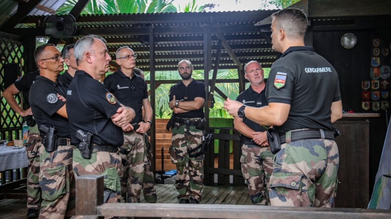 Dans un abri au sol en bois, toit en tôle et sans mur, le DGGN, au premier plan, ainsi que le COMGEND et le CGOM, à côté de lui, écoutent un gendarme, de trois quarts dos, à droite.