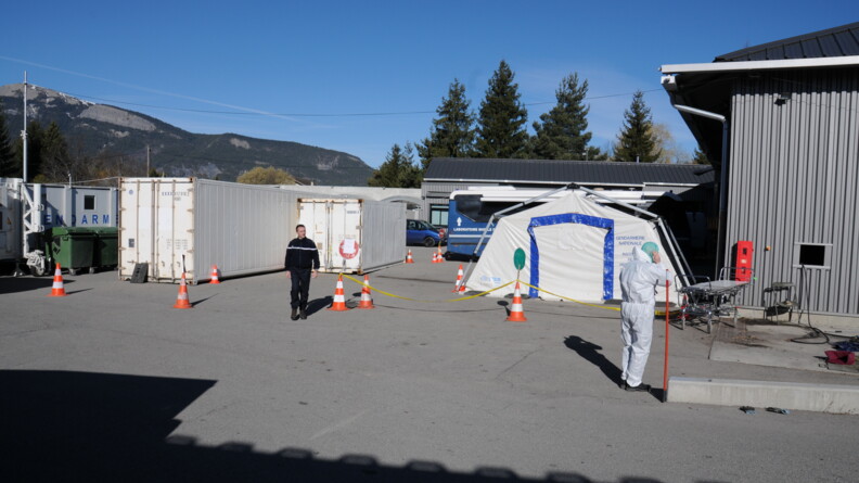 Un gendarme marche devant un hangar et une tente de l'IRCGN. Derri&egrave;re lui se trouve le bus de l'IRCGN