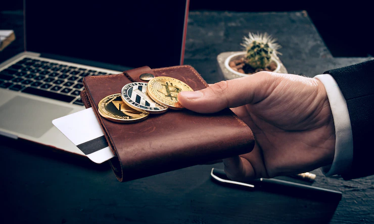 Agrandir l'image Photo d'illustration représentant un homme tendant un portefeuille avec des grandes pièces de monnaie, devant un bureau sur lequel est posé un ordinateur.
